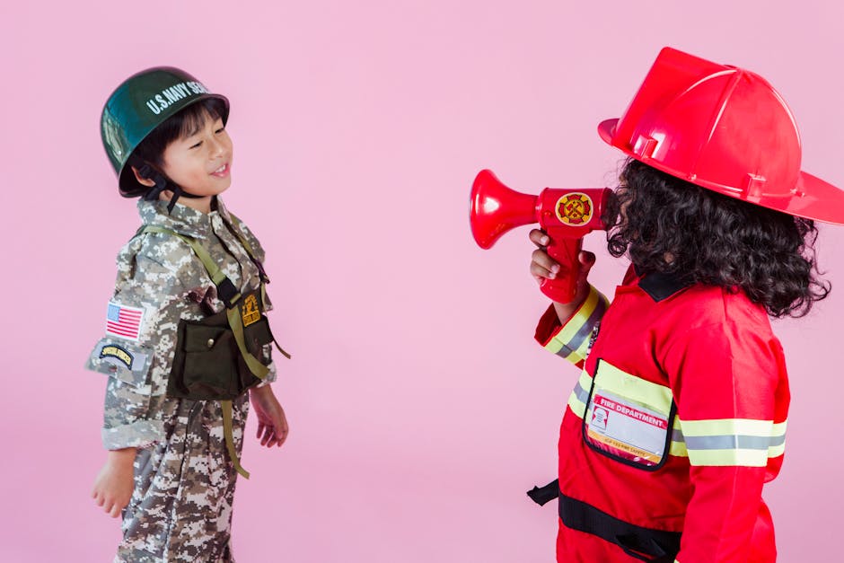 Side view of multiethnic kids wearing navy uniform and firefighter costume with loudspeaker standing together on pink background in hardhats and looking at each other